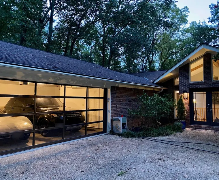 A full-view glass and black aluminum garage door lit from inside at dusk, on a brick house among tall trees