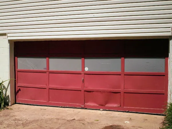 A red garage door with a dented bottom panel and covered windows
