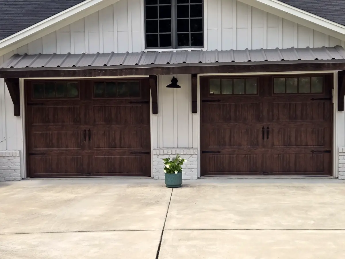 Two dark wood-look carriage garage doors under a metal awning on a white board-and-batten house
