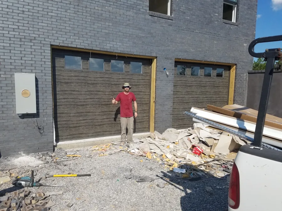 A new dark wood-look garage door with a row of windows on a dark brick house under construction, a man giving a thumbs-up in front