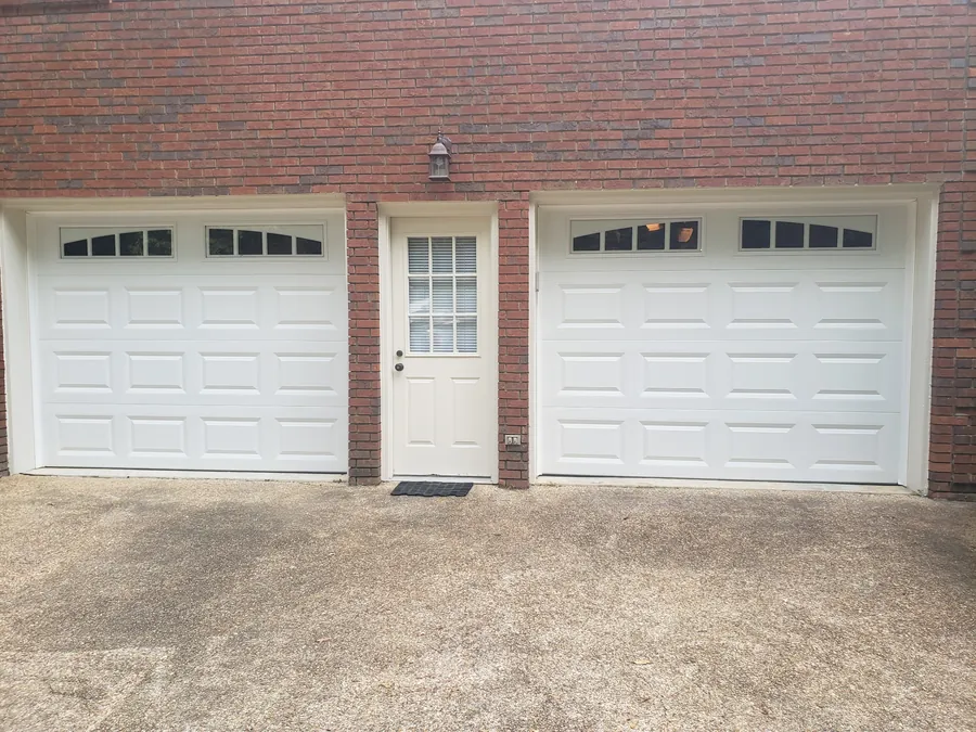 Two new white raised-panel garage doors with arched windows on a red brick house
